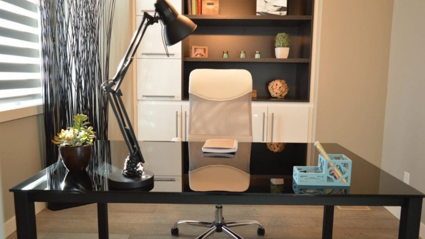 Imagem de um escritório, há uma prateleira ao fundo, uma cadeira branca e uma mesa preta, sobre ela há um organizador, um vaso uma luminária e um caderno